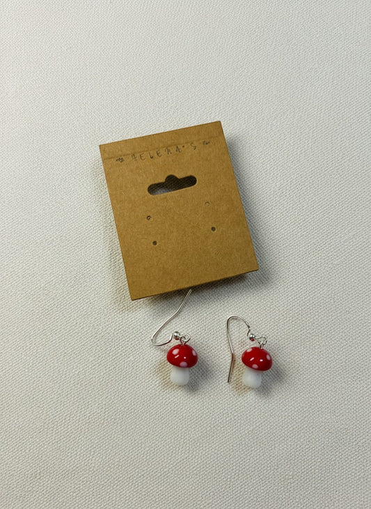 Helena’s Artisan & Gift Co. handcrafted red and white glass mushroom dangle earrings on a brown display card.